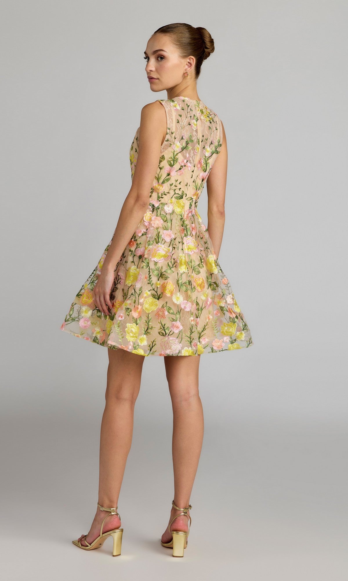 Model in profile gazes over her shoulder, showcasing the Mac Duggal 56409 Short Party Dress—a sleeveless yellow floral lace mini dress with a flattering fit-and-flare silhouette—paired with beige heels against a plain background.